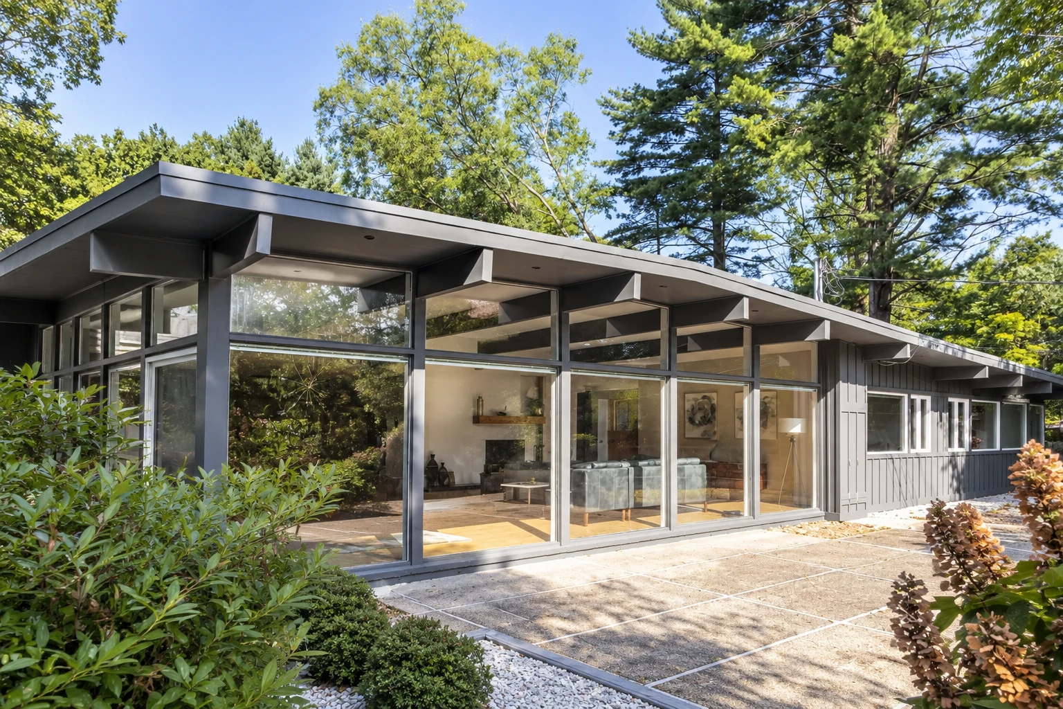 Modern all-season room with dark framing, full-height glass walls, and a low sloped roof beside a wooded patio.
