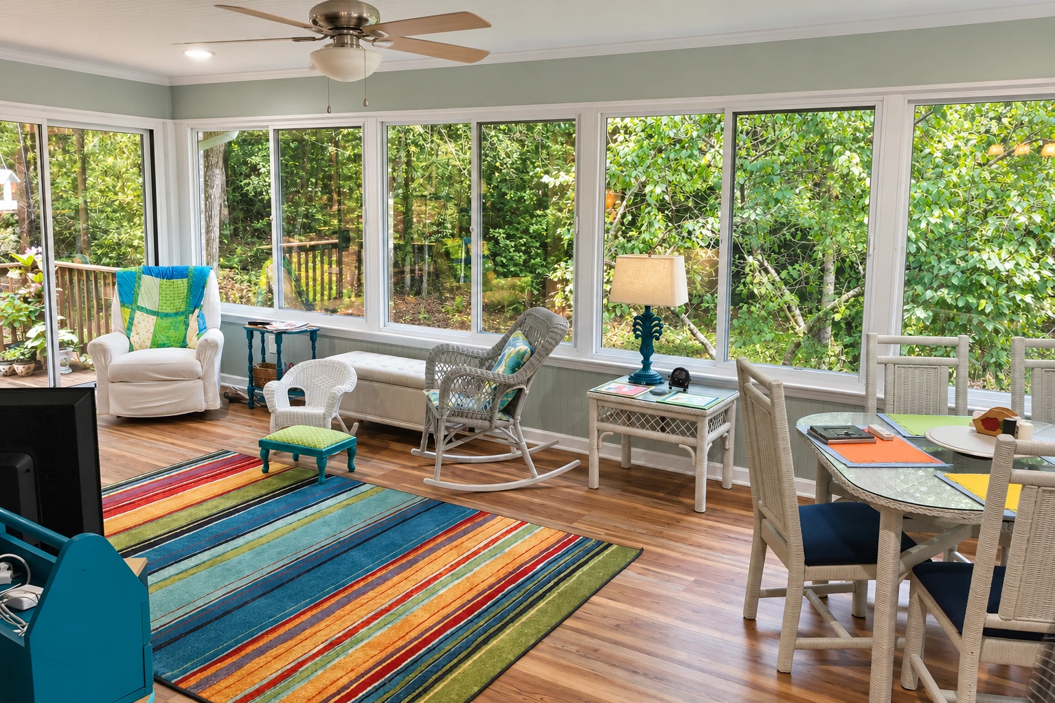 Converted deck sunroom furnished with wicker seating, a dining table, and wide windows facing leafy trees.