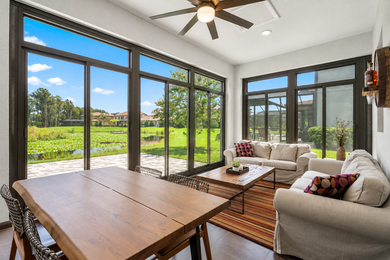 Bright four-season sunroom with dark-framed windows, a dining table, and sofas overlooking a green lawn and pond.