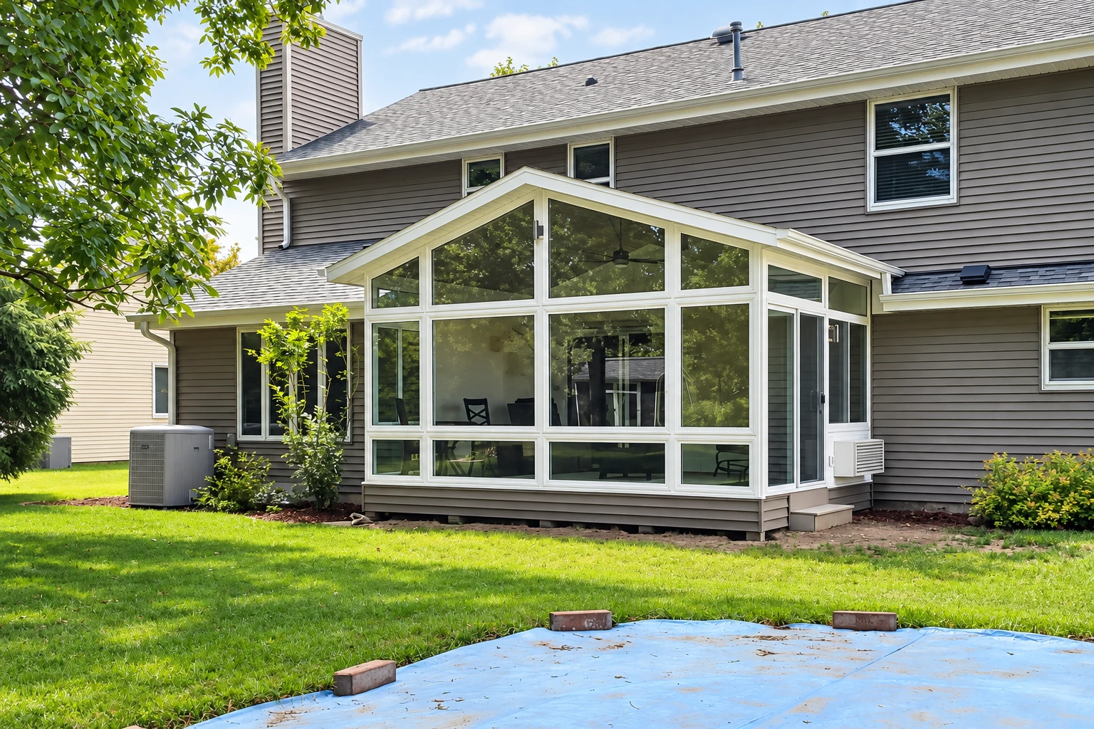Enclosed patio sunroom with a peaked roof and white-framed windows, attached to a gray-sided house.