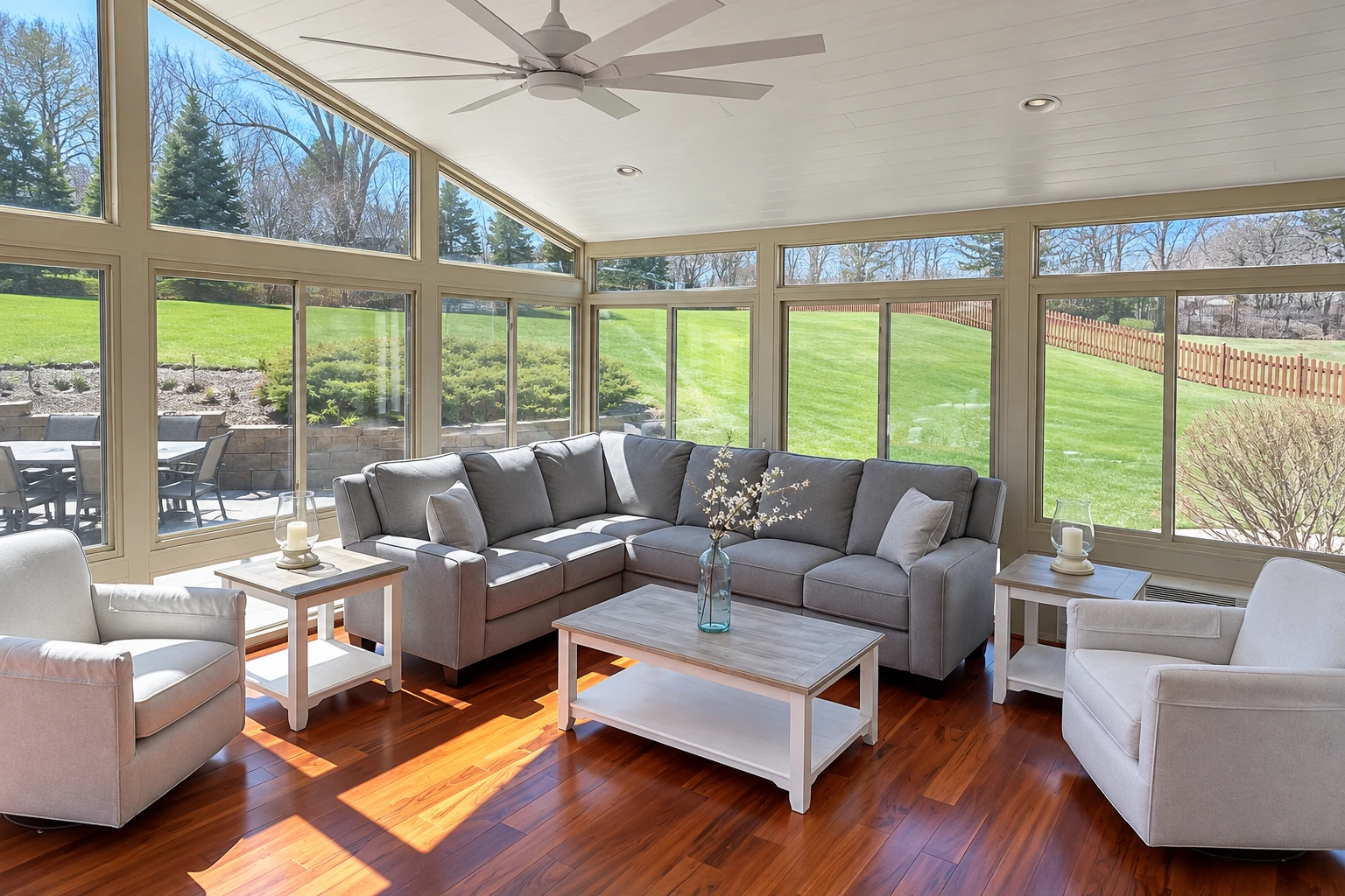 Sunroom with broad windows, a gray sectional, and wood floors overlooking a lawn and patio.