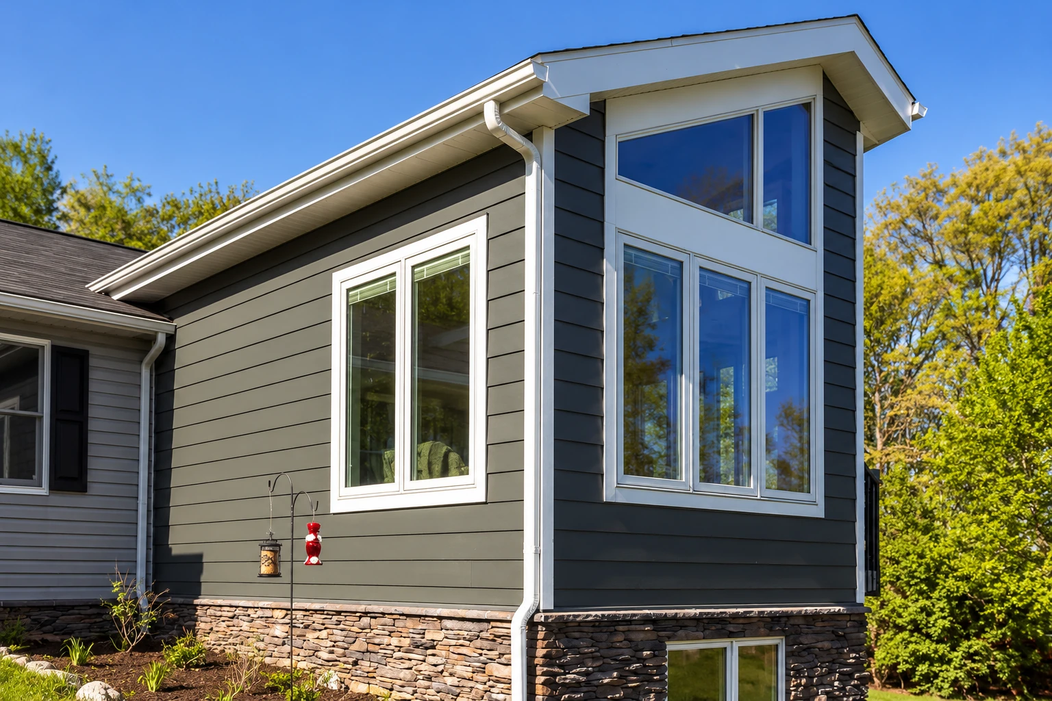 Sunroom addition with dark horizontal siding, tall white-trimmed windows, and a stone foundation beneath a clear blue sky.