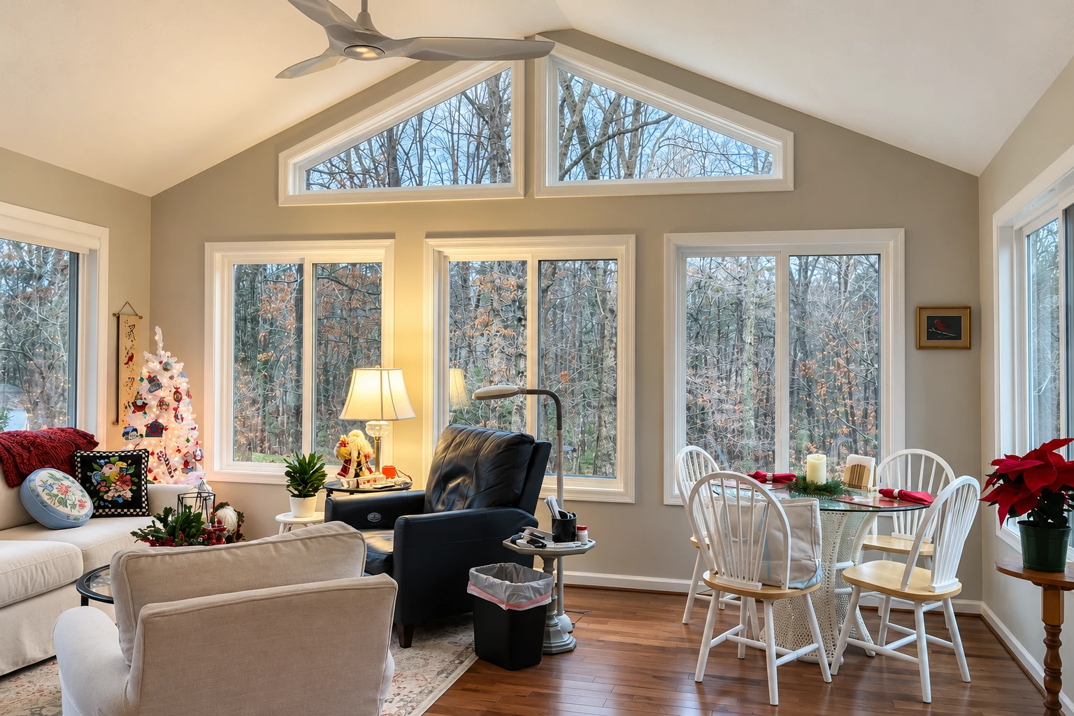 Furnished sunroom with a vaulted ceiling, tall windows overlooking bare trees, and a dining area set for the holidays.