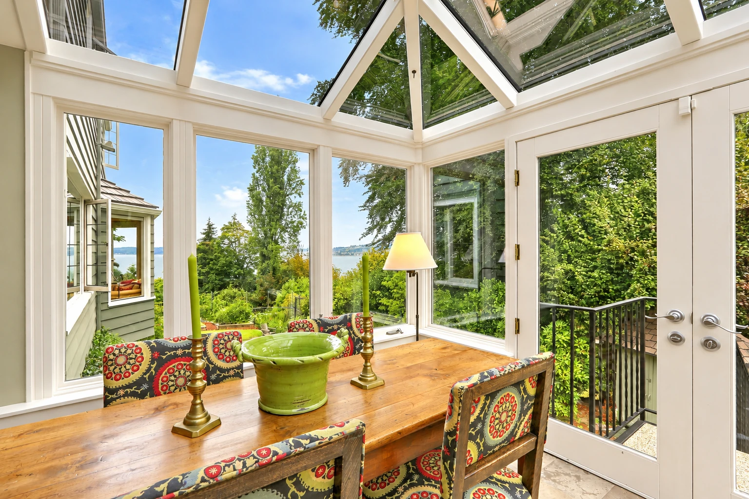 Sunroom with a glass roof and walls, furnished with a long wooden table and colorful patterned chairs.