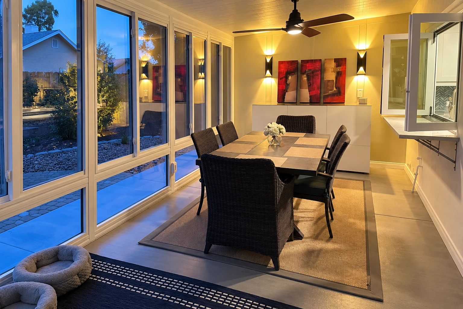Three-season sunroom with a dining table, dark wicker chairs, and tall windows overlooking a garden at dusk.
